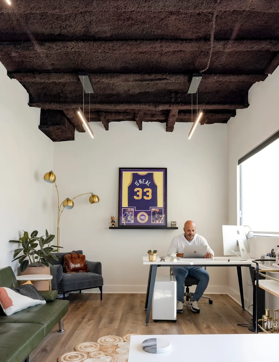 The Rigs Workspace private office with framed Shaq O'Neal jersey, exposed dark ceiling beams, brass floor lamp, and member at dual-monitor desk