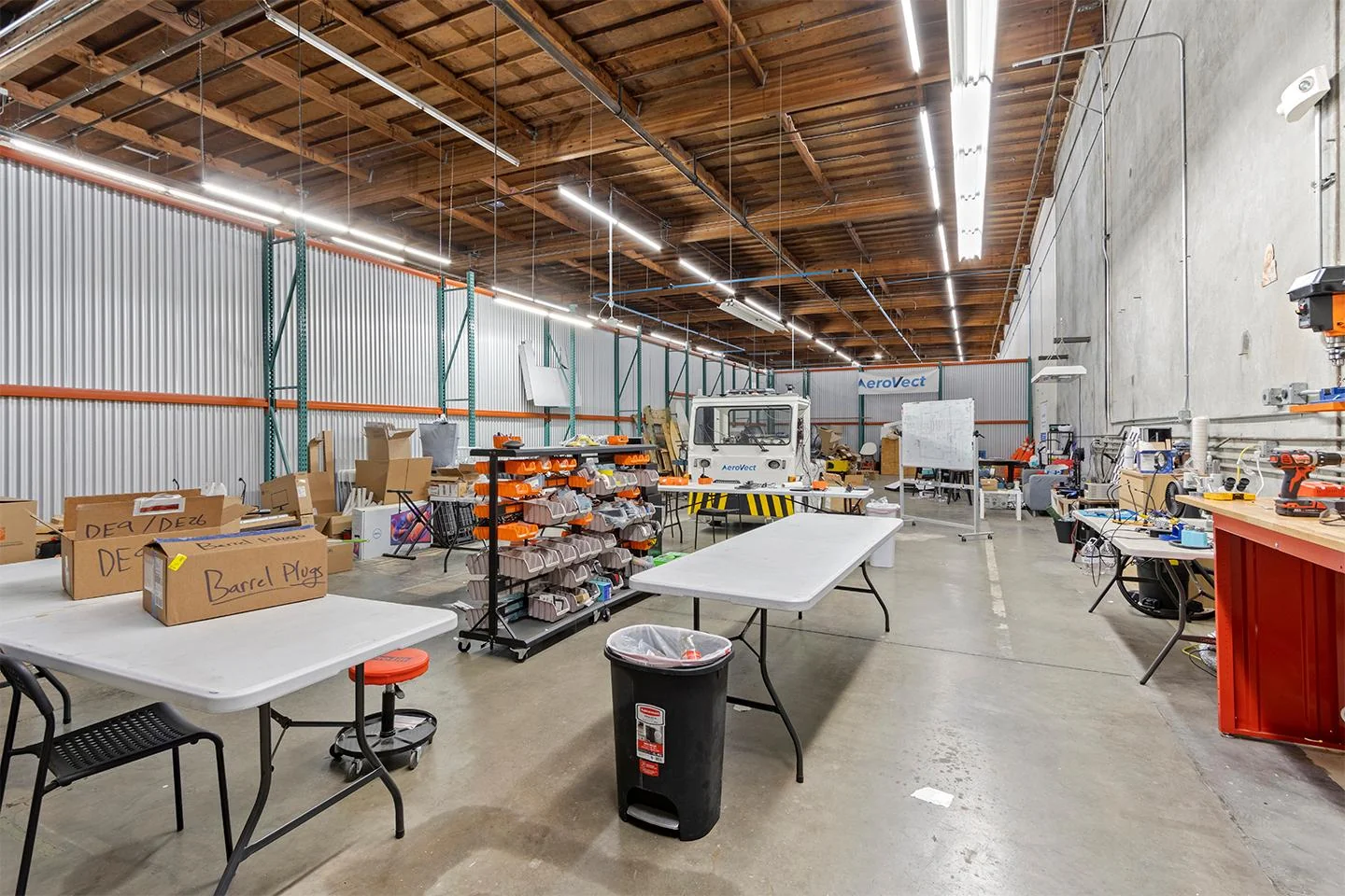 ReadySpaces industrial warehouse with exposed wood-beam ceiling, corrugated metal walls, pallet racking, AeroVect autonomous vehicle, folding tables, and parts bins