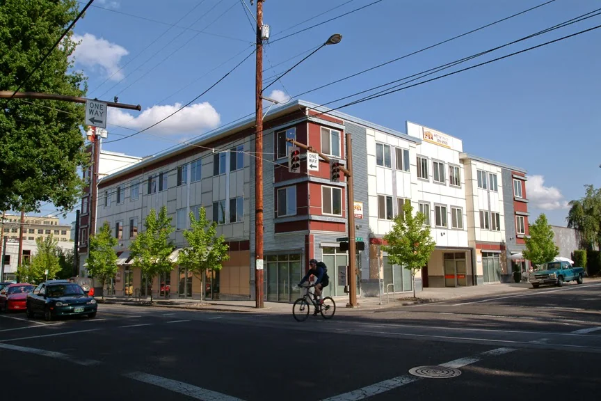 ActivSpace on Lovejoy modern three-story mixed-use building with gray and burgundy metal cladding, street-level storefronts, cyclist passing, and green trees