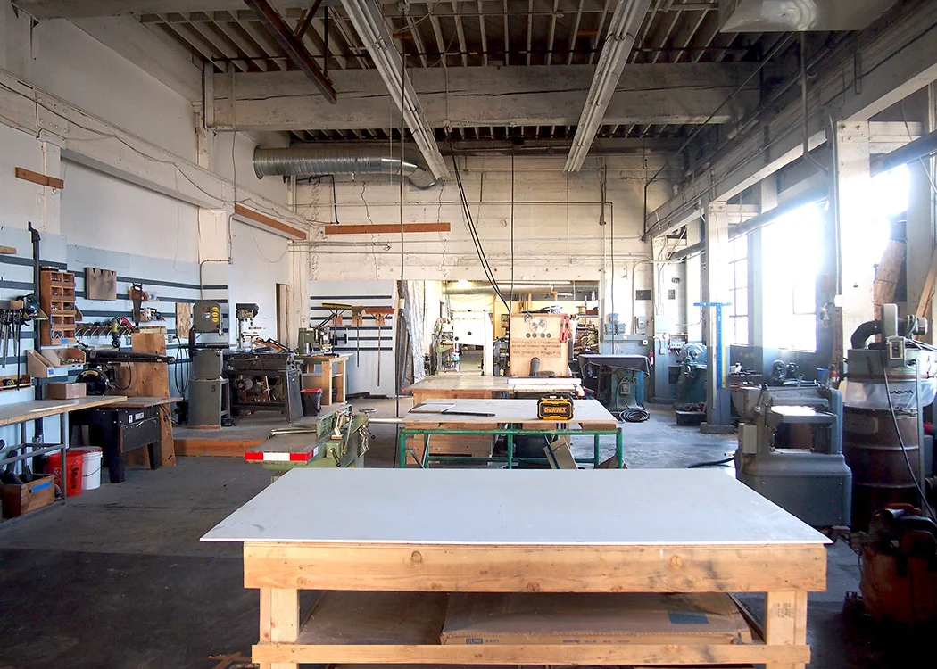 Past Lives Makerspace industrial woodworking shop with large work tables, band saws, drill presses, power tools, high ceilings with exposed beams, and natural light