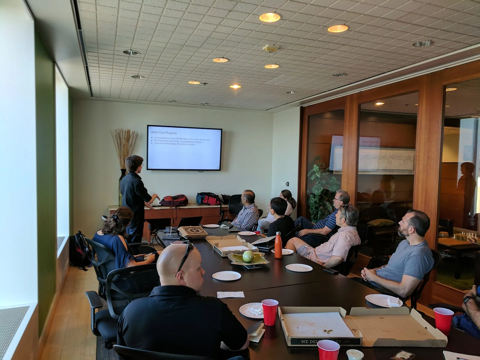 NedSpace conference room with presenter giving a talk at wall-mounted screen, attendees seated around dark table, wood-framed glass walls, and warm lighting