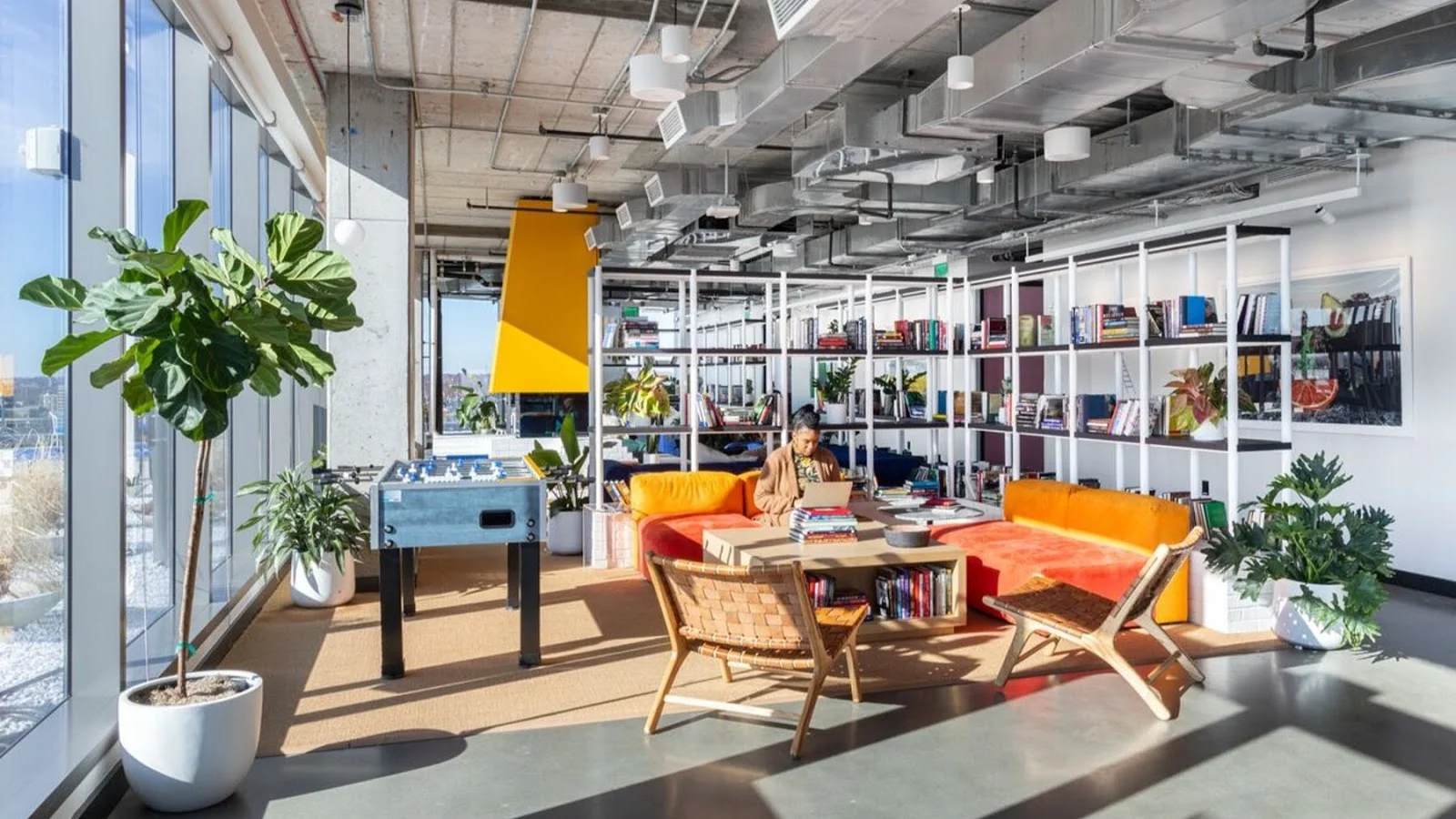 WeWork vibrant lounge with orange and yellow curved sofa, foosball table, bookshelves, large plants, and natural light